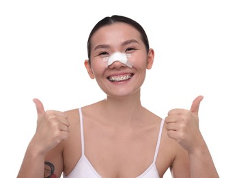 Photo of Woman with medical bandage on her nose after plastic surgery operation showing thumbs up against white background