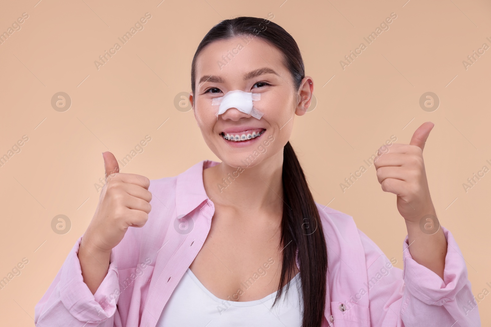 Photo of Woman with medical bandage on her nose after plastic surgery operation showing thumbs up against beige background