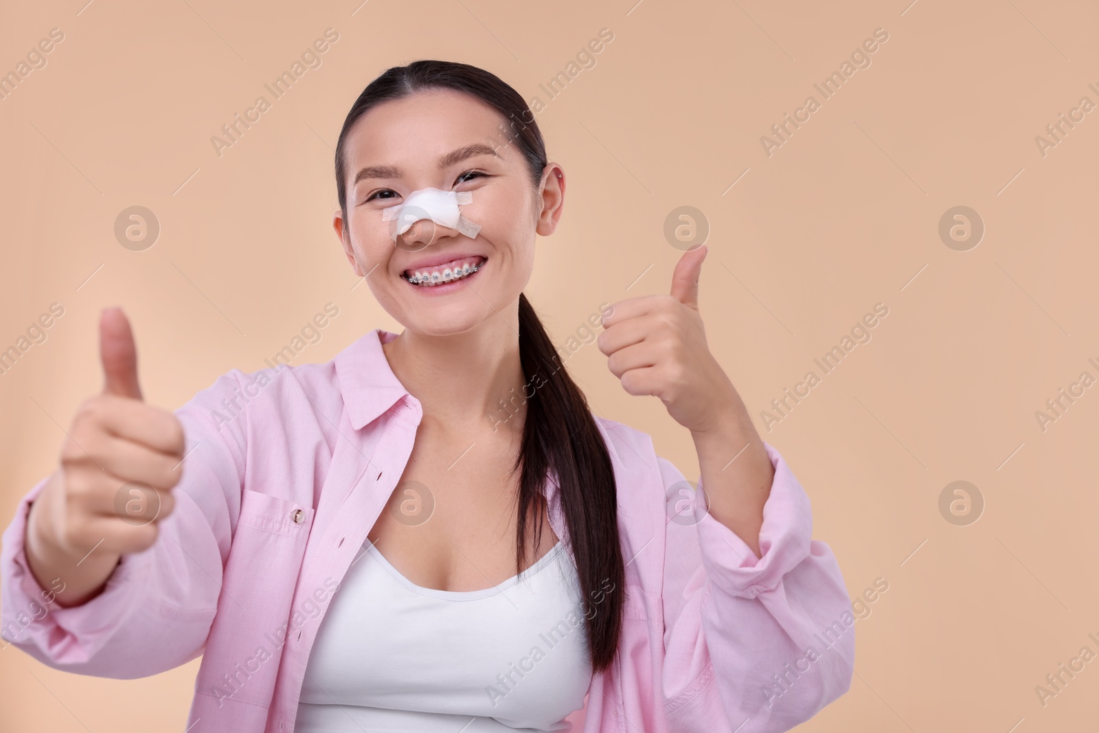 Photo of Woman with medical bandage on her nose after plastic surgery operation showing thumbs up against beige background