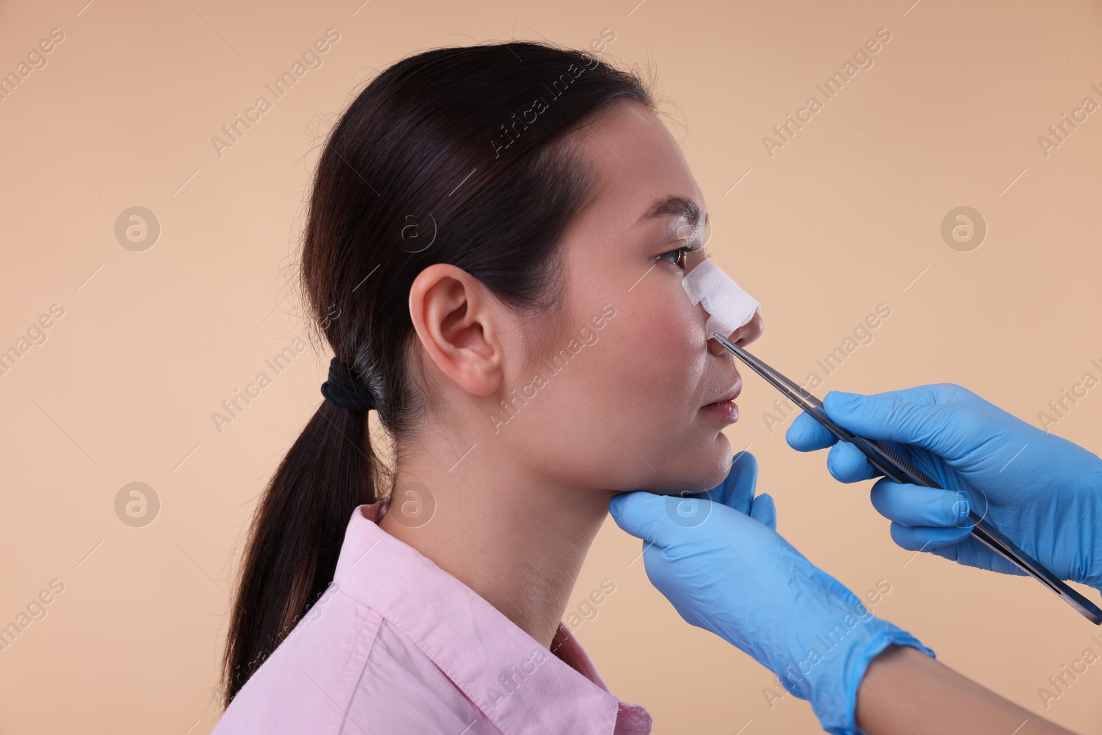 Photo of Doctor removing medical bandage from patient's nose after plastic surgery operation on beige background, closeup