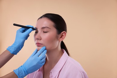 Doctor drawing marks on patient's nose for plastic surgery operation against beige background, closeup. Space for text