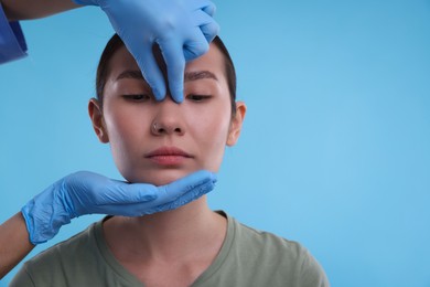 Photo of Doctor checking patient's nose before plastic surgery operation on light blue background, closeup