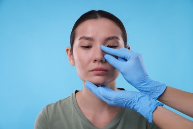 Photo of Doctor checking patient's nose before plastic surgery operation on light blue background, closeup