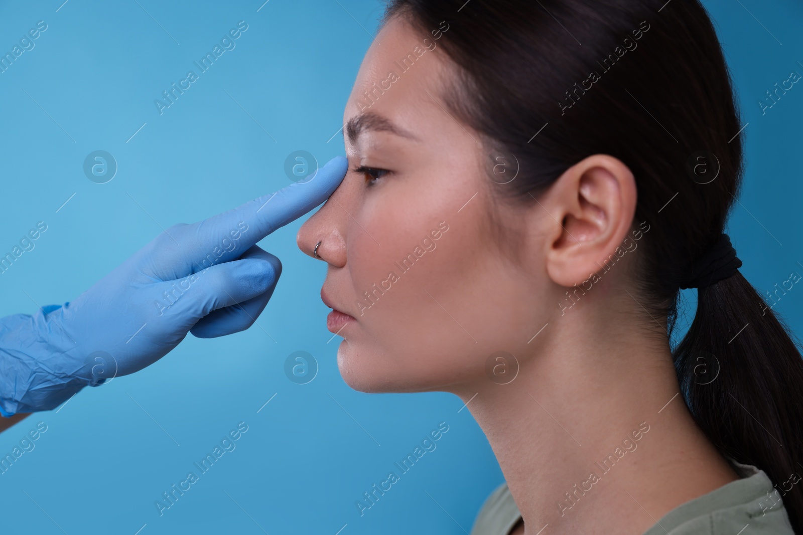 Photo of Doctor checking patient's nose before plastic surgery operation on light blue background, closeup