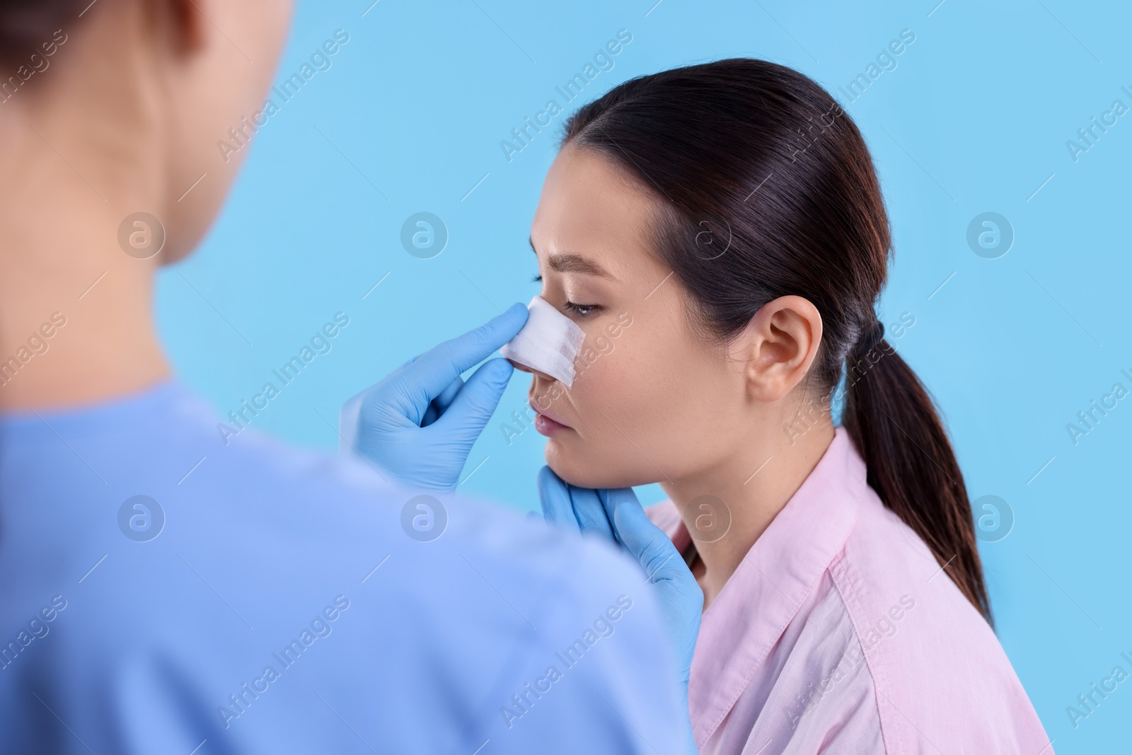 Photo of Doctor checking patient's nose after plastic surgery operation on light blue background, closeup