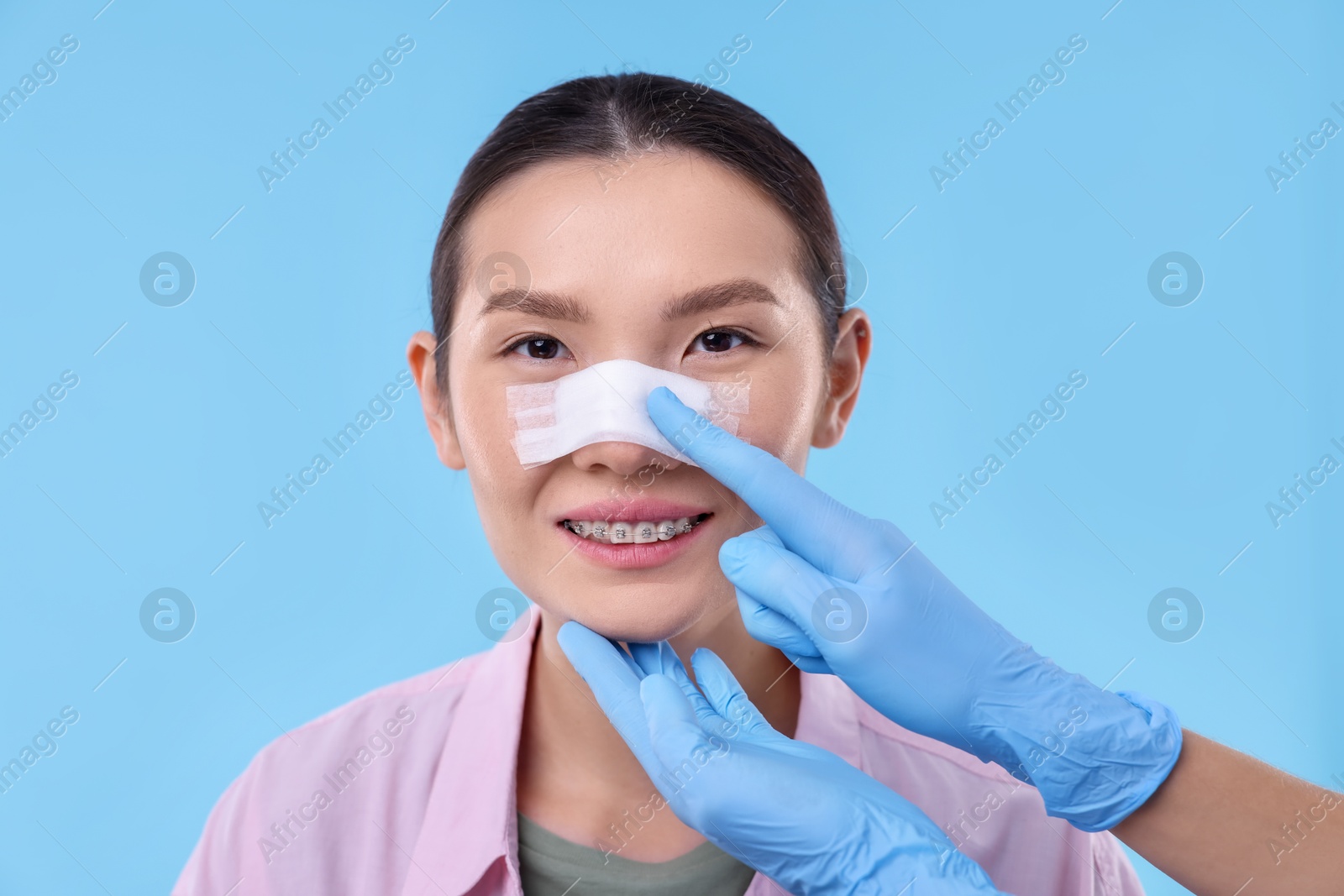 Photo of Doctor checking patient's nose after plastic surgery operation on light blue background, closeup