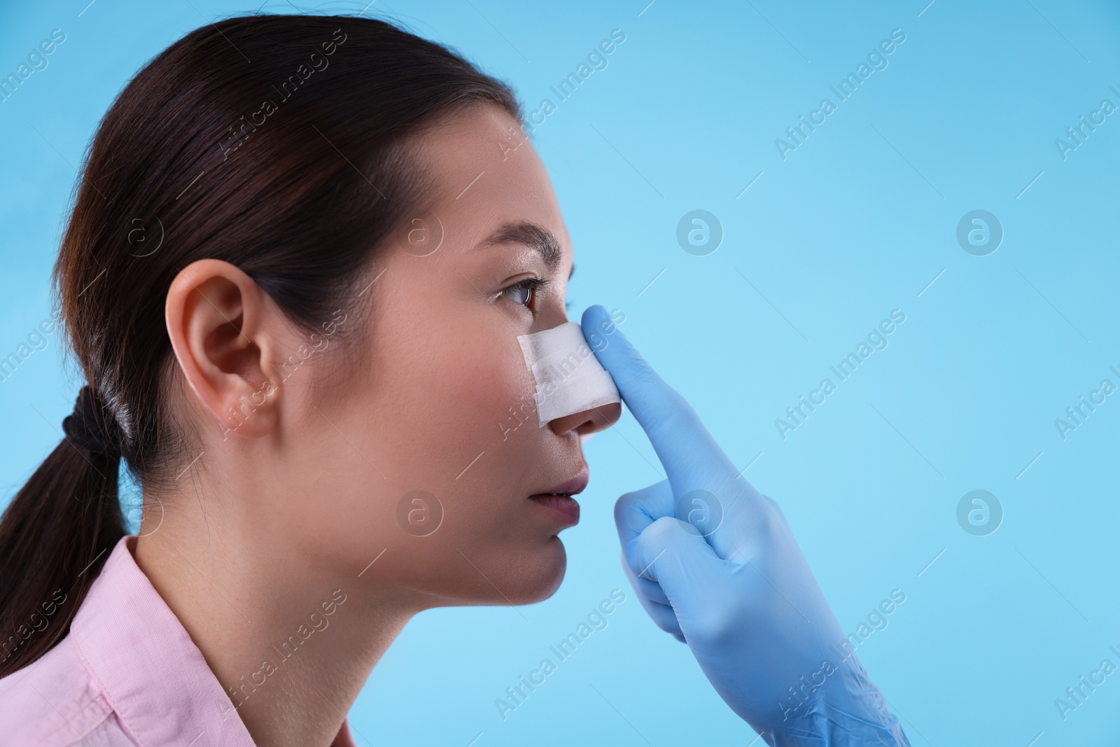 Photo of Doctor checking patient's nose after plastic surgery operation on light blue background, closeup