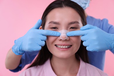 Photo of Doctor checking patient's nose after plastic surgery operation on pink background, closeup