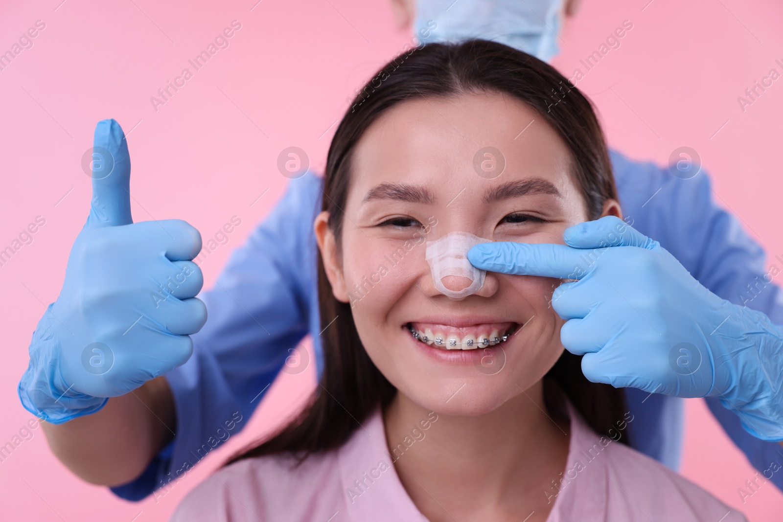 Photo of Doctor checking patient's nose after plastic surgery operation and showing thumbs up on pink background, closeup