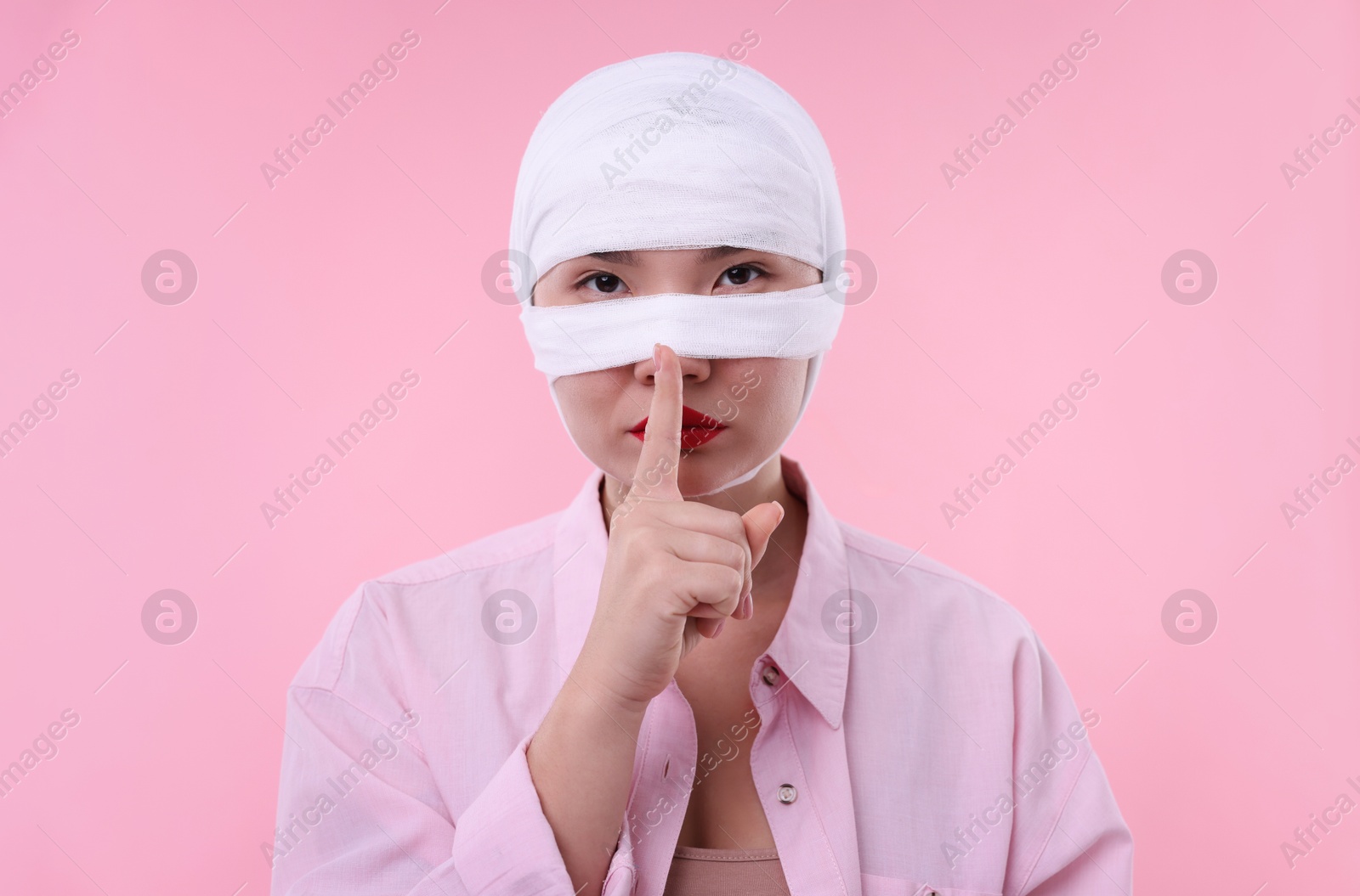 Photo of Woman with nose wrapped in medical bandage after plastic surgery operation making silent gesture on pink background