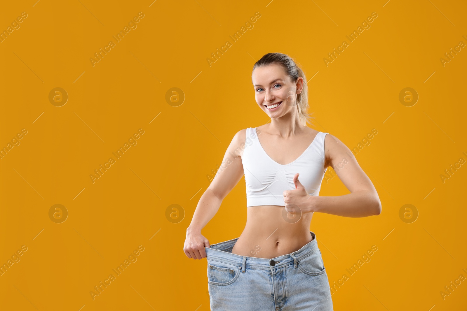 Photo of Weight loss. Happy woman wearing big jeans and showing thumbs up on yellow background, space for text