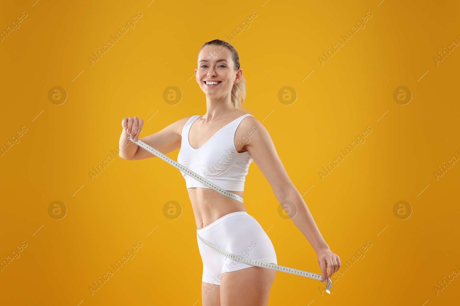 Photo of Weight loss. Happy woman measuring her slim waist with tape on yellow background