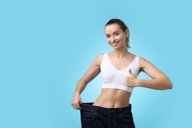 Photo of Weight loss. Happy woman wearing big jeans and showing thumbs up on light blue background. Space for text