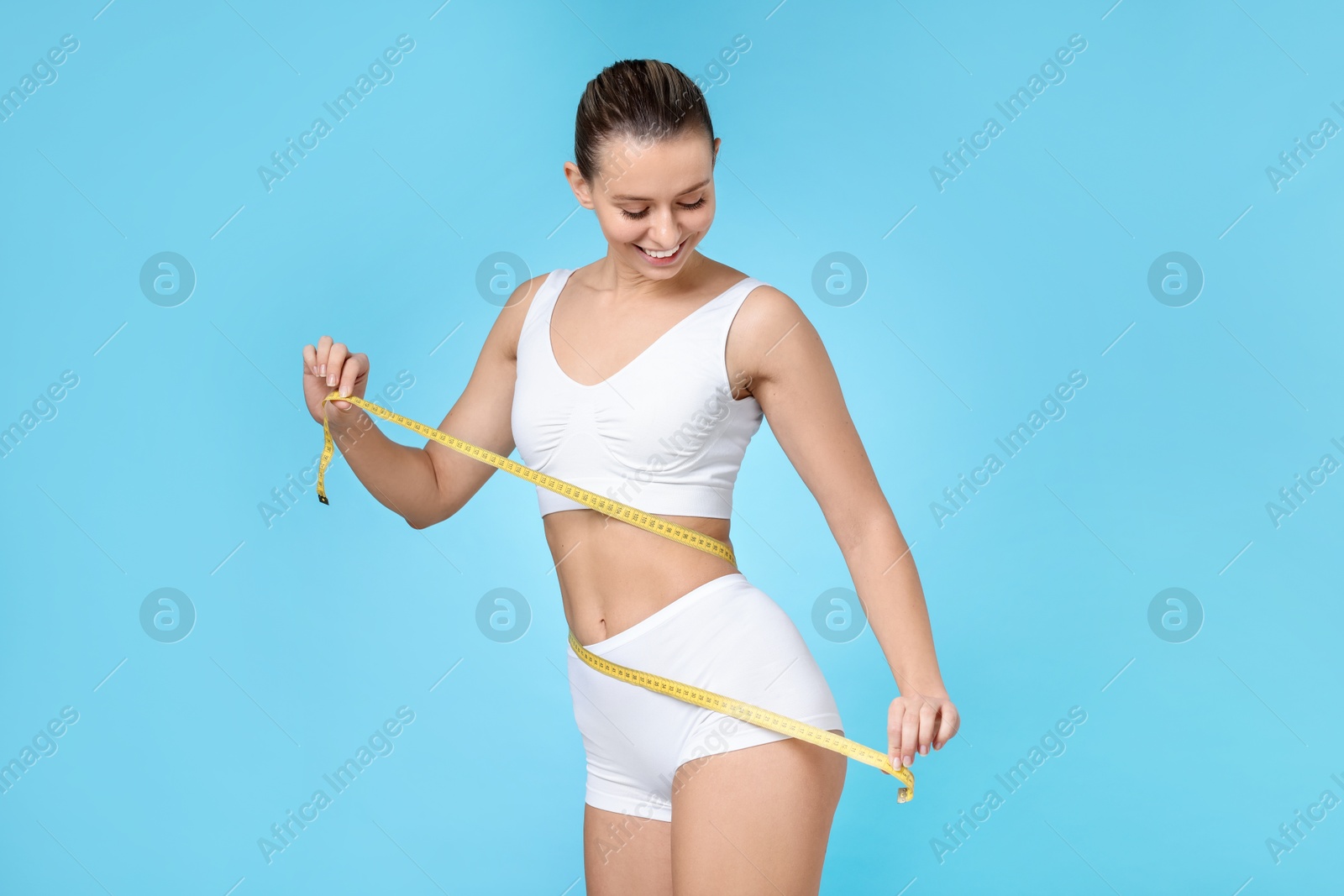 Photo of Weight loss. Happy woman measuring her slim waist with tape on light blue background