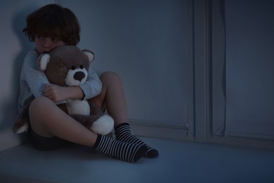 Photo of Fearful boy hiding with teddy bear on windowsill at night. Space for text