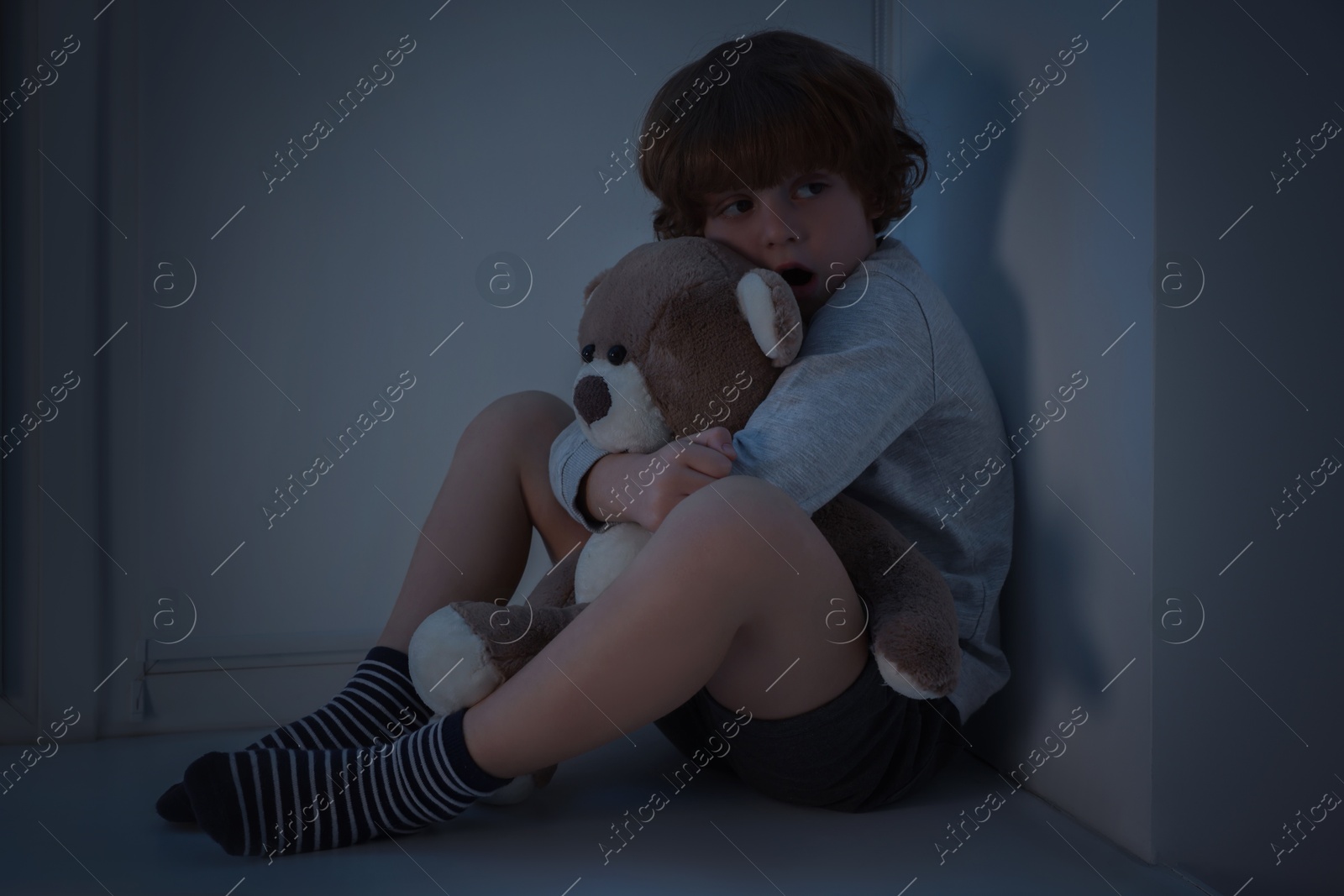 Photo of Fearful boy hiding with teddy bear on windowsill at night