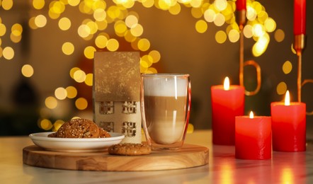 Photo of Aromatic coffee in glass, tasty cookies and burning candles on table against blurred Christmas lights. Bokeh effect