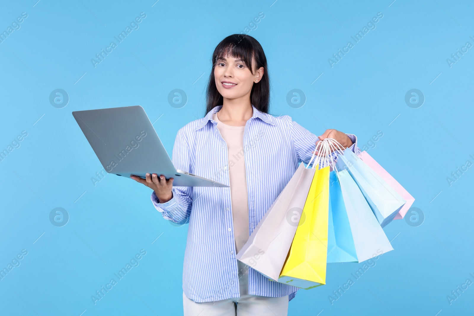 Photo of Internet shopping. Happy woman with laptop and colorful bags on light blue background