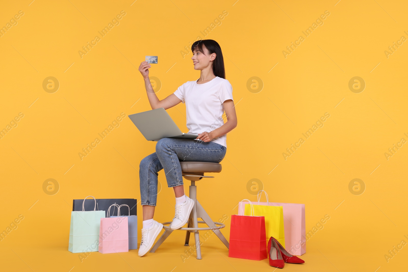 Photo of Internet shopping. Happy woman with credit card and laptop sitting on stool among colorful bags against orange background