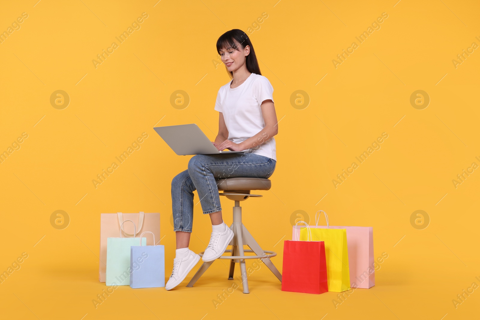 Photo of Internet shopping. Happy woman with laptop sitting on stool among colorful bags against orange background