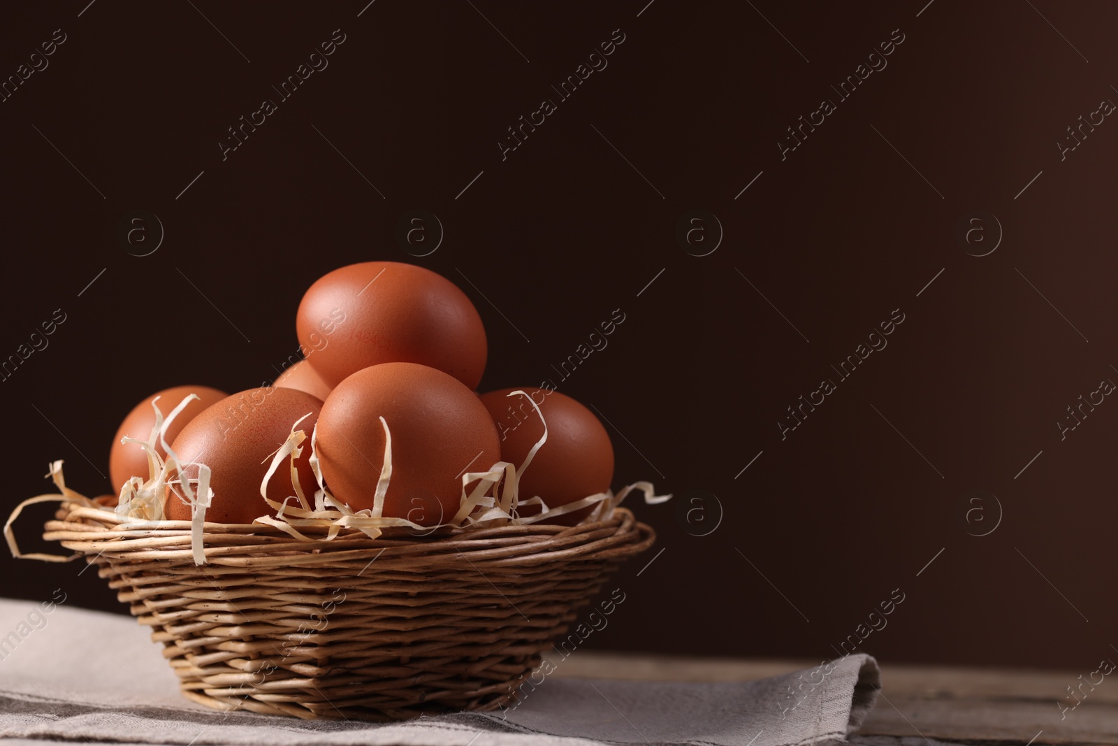 Photo of Wicker basket with fresh eggs on wooden table against brown background, closeup. Space for text