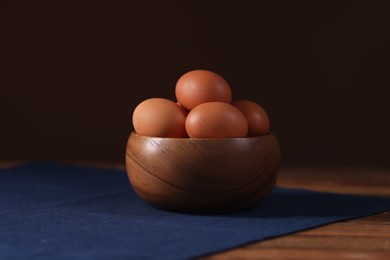 Photo of Fresh eggs in bowl on wooden table against brown background, closeup