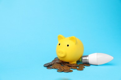 Photo of Piggy bank, coins and lightbulb on light blue background, space for text. Energy saving concept