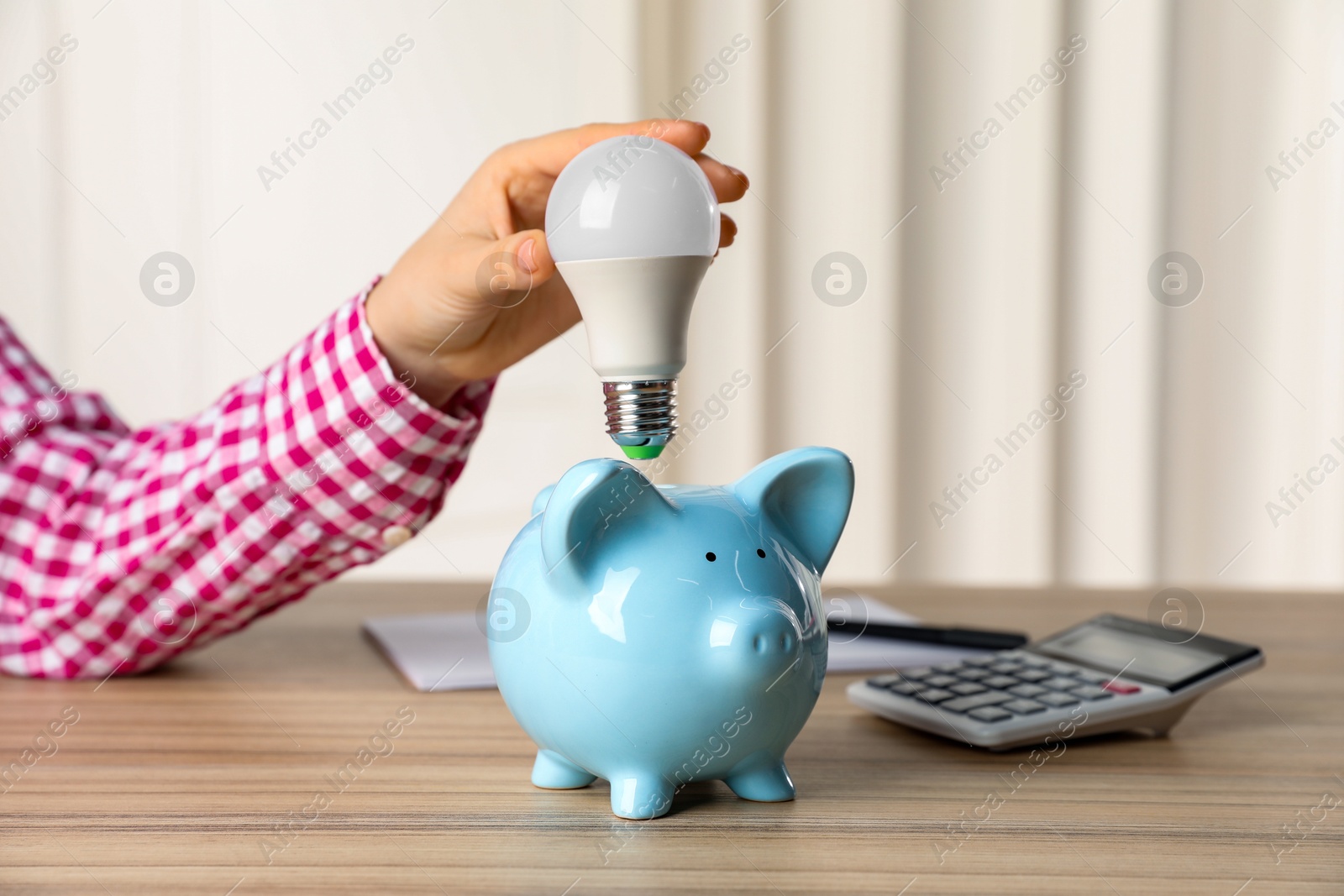 Photo of Woman holding light bulb above piggy bank at wooden table, closeup. Energy saving concept