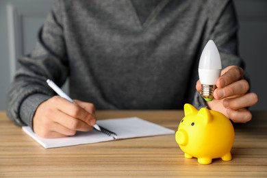 Photo of Man holding light bulb above piggy bank while taking notes at wooden table, closeup. Energy saving concept