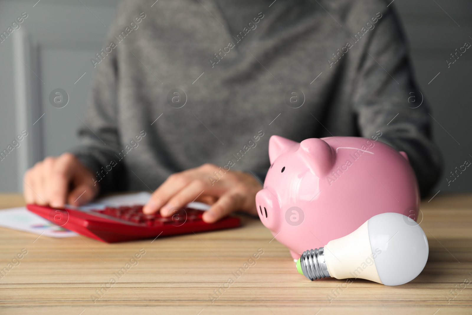 Photo of Man using calculator at wooden table, focus on piggy bank and light bulb. Energy saving concept