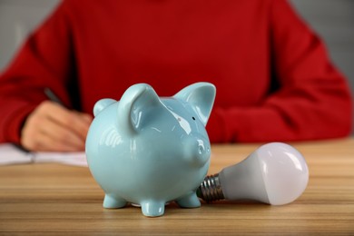 Photo of Man taking notes at wooden table, focus on piggy bank and light bulb. Energy saving concept