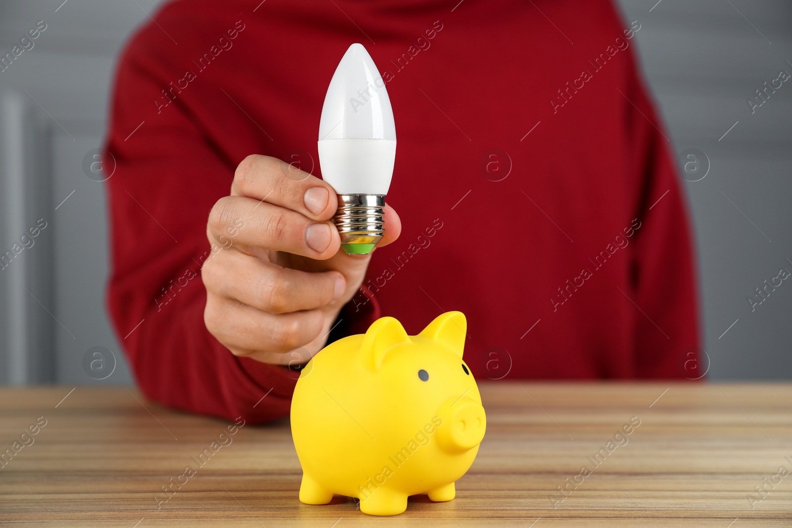 Photo of Man holding light bulb above piggy bank at wooden table, closeup. Energy saving concept