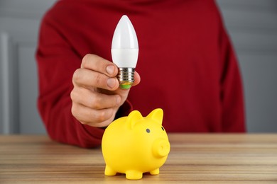 Photo of Man holding light bulb above piggy bank at wooden table, closeup. Energy saving concept