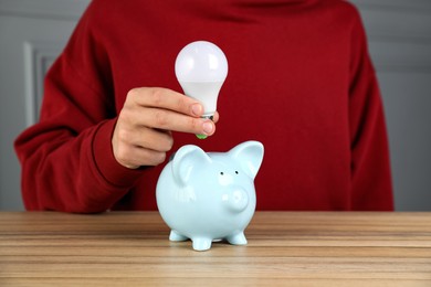 Photo of Man holding light bulb above piggy bank at wooden table, closeup. Energy saving concept