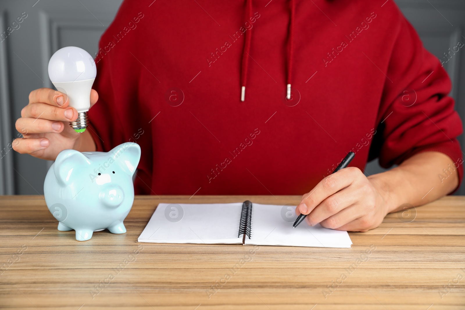 Photo of Man holding light bulb above piggy bank while taking notes at wooden table, closeup. Energy saving concept