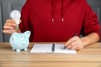 Man holding light bulb above piggy bank while taking notes at wooden table, closeup. Energy saving concept