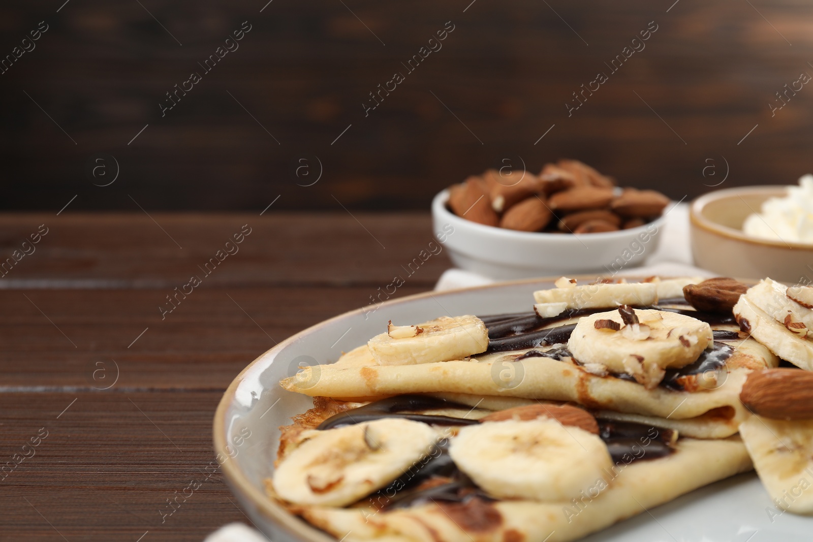 Photo of Delicious crepes with chocolate sauce, banana and almonds served on wooden table, closeup. Space for text