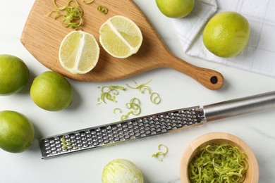 Photo of Lime zest, fresh fruits and grater on white marble table, flat lay