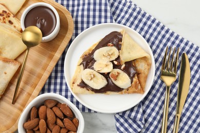 Photo of Delicious crepes with chocolate sauce, banana and almonds served on white table, flat lay