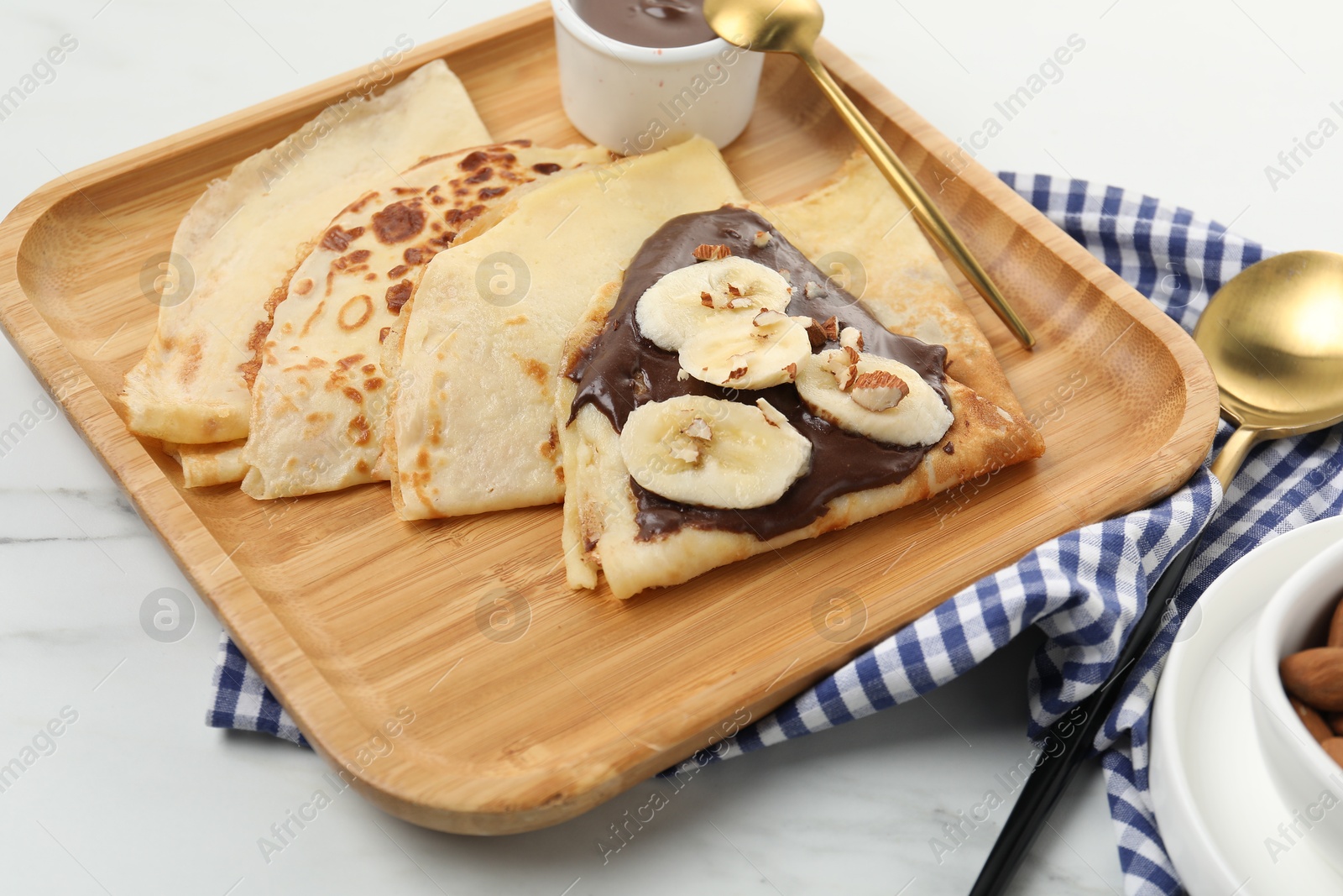 Photo of Delicious crepes with chocolate sauce, banana and almonds served on white marble table, closeup