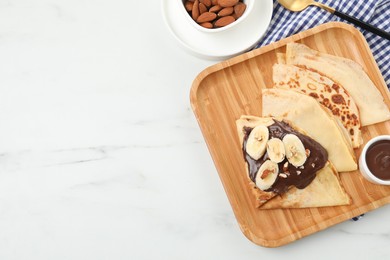 Photo of Delicious crepes with chocolate sauce, banana and almonds on white marble table, top view. Space for text