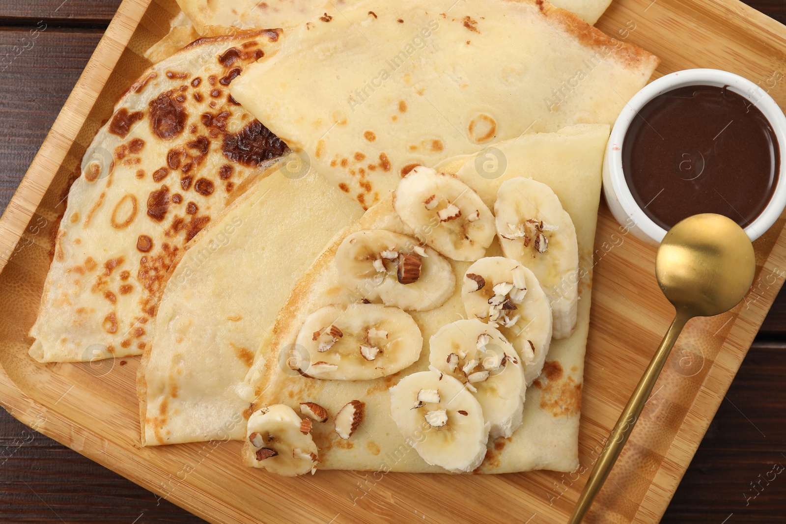 Photo of Delicious crepes with chocolate sauce, banana and almonds on wooden table, top view