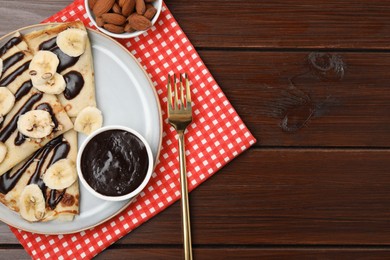 Photo of Delicious crepes with chocolate sauce, banana and almonds served on wooden table, flat lay. Space for text