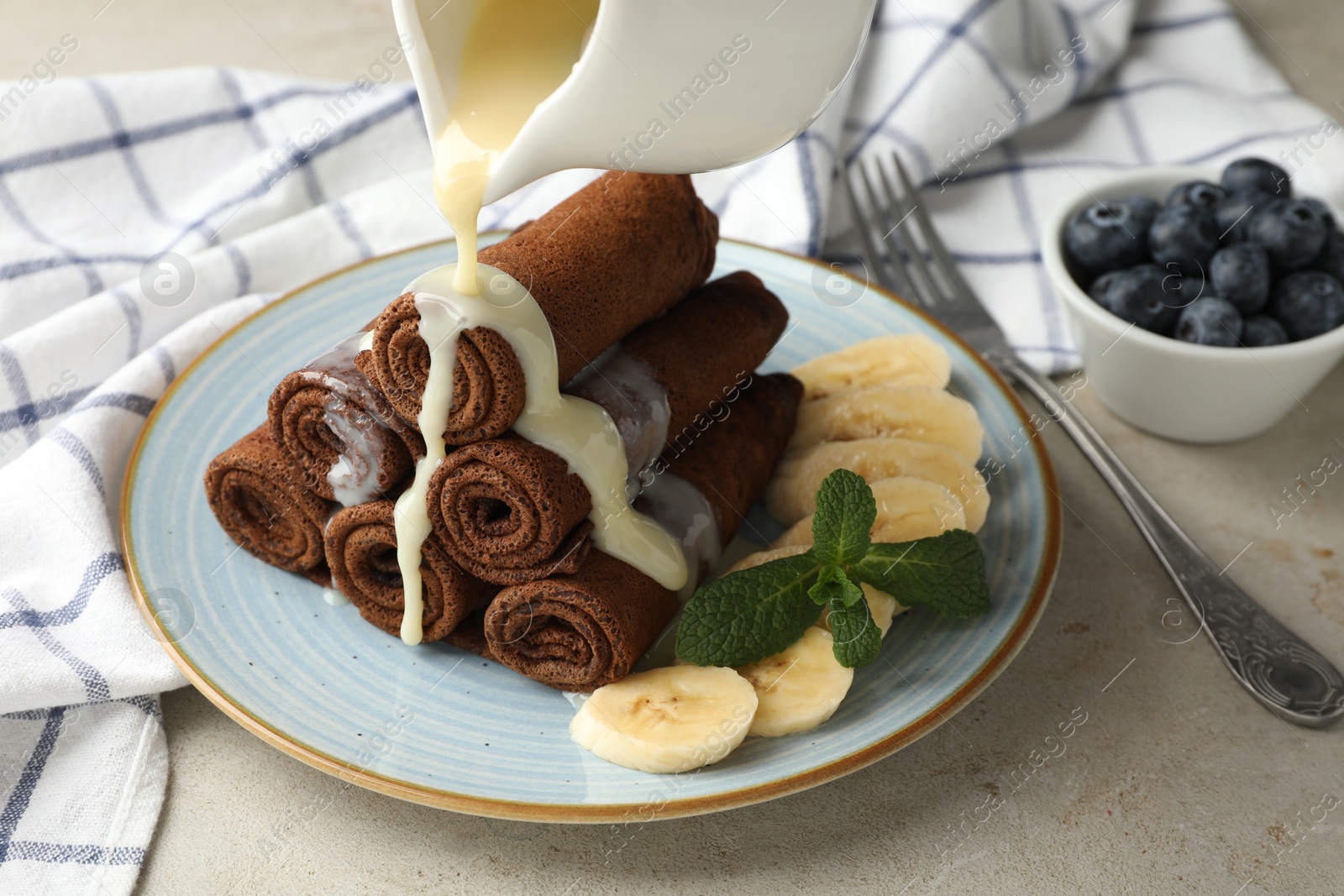 Photo of Pouring condensed milk onto chocolate crepes with banana on light table, closeup