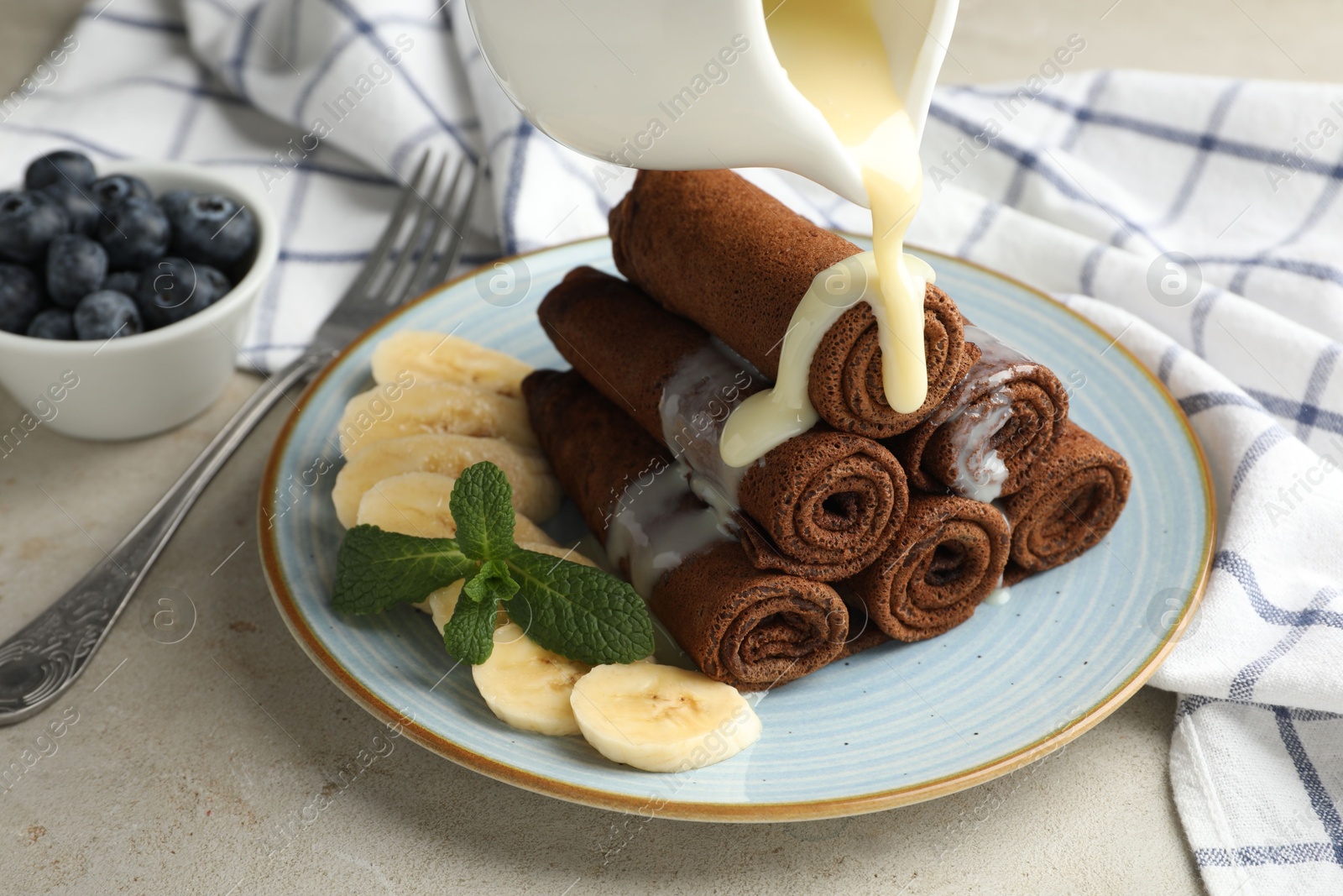 Photo of Pouring condensed milk onto chocolate crepes with banana on light table, closeup