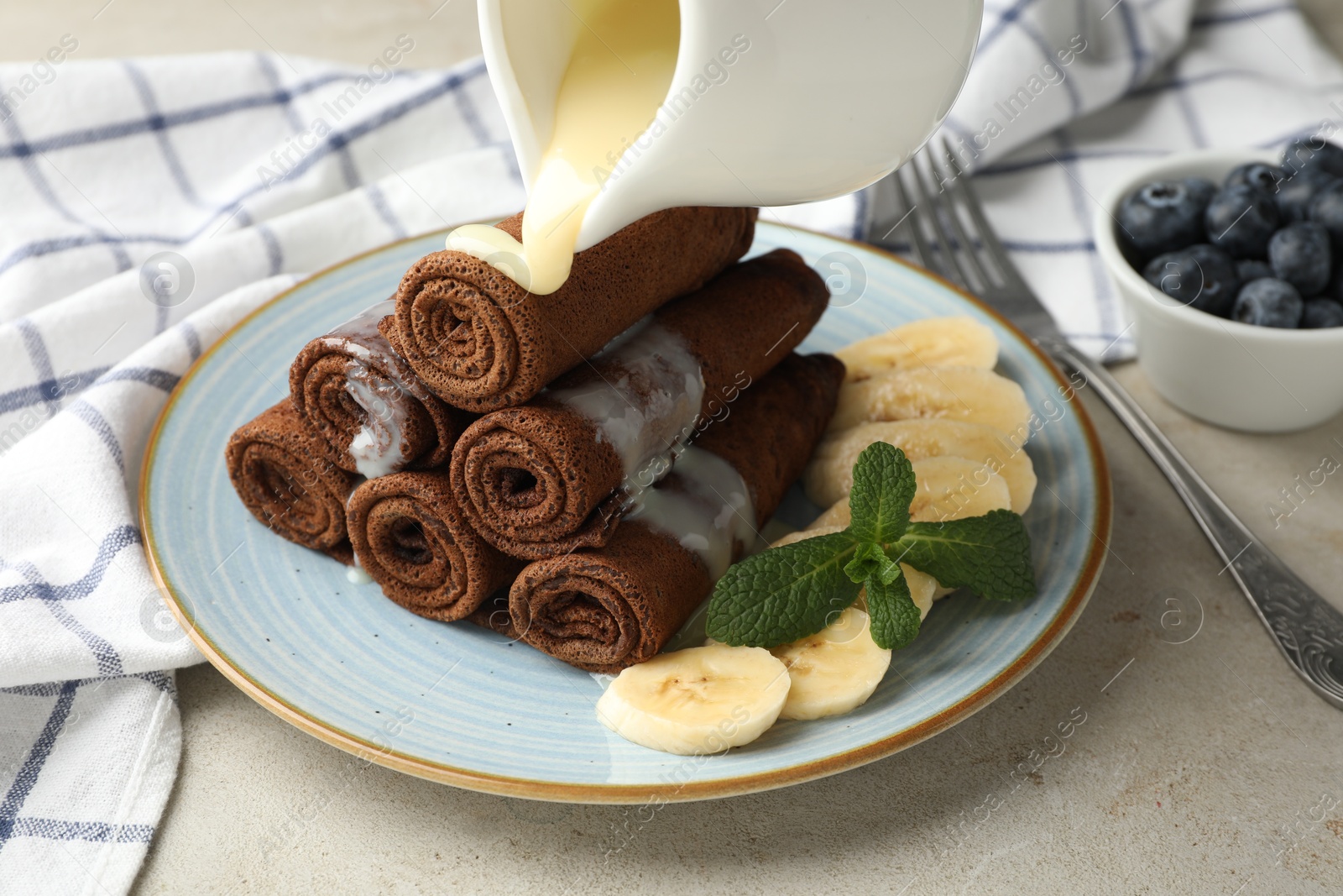 Photo of Pouring condensed milk onto chocolate crepes with banana on light table, closeup