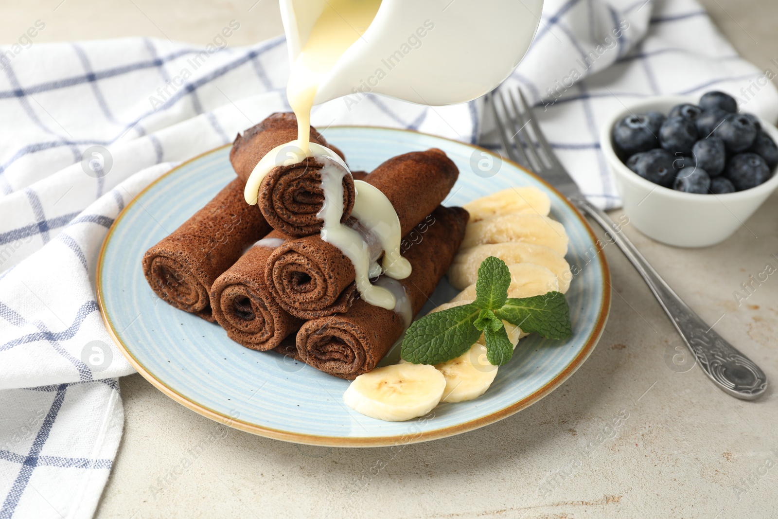 Photo of Pouring condensed milk onto chocolate crepes with banana on light table, closeup