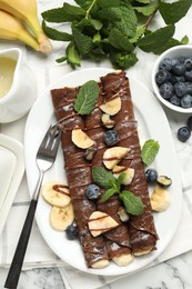 Photo of Delicious chocolate crepes with banana and blueberries served on white marble table, flat lay