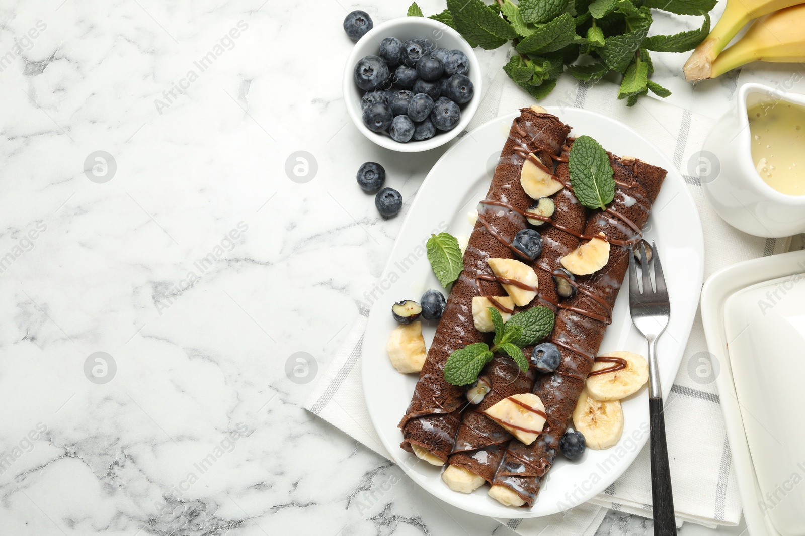 Photo of Delicious chocolate crepes with banana and blueberries served on white marble table, flat lay. Space for text