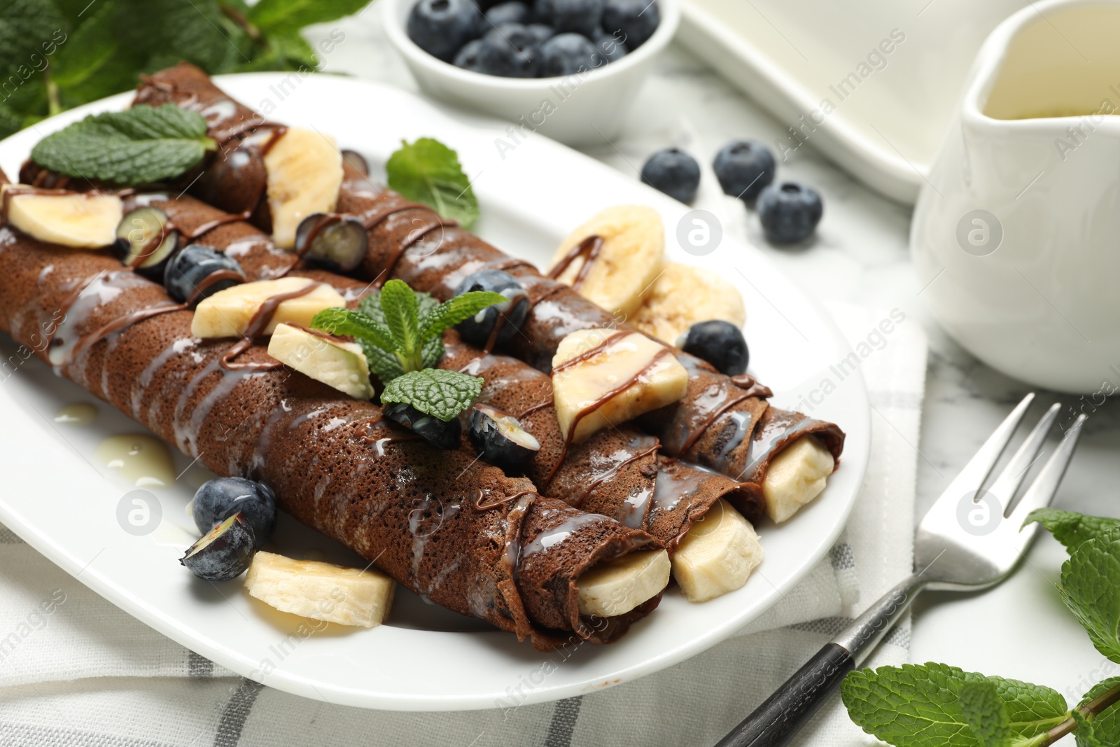 Photo of Delicious chocolate crepes with banana and blueberries served on white marble table, closeup